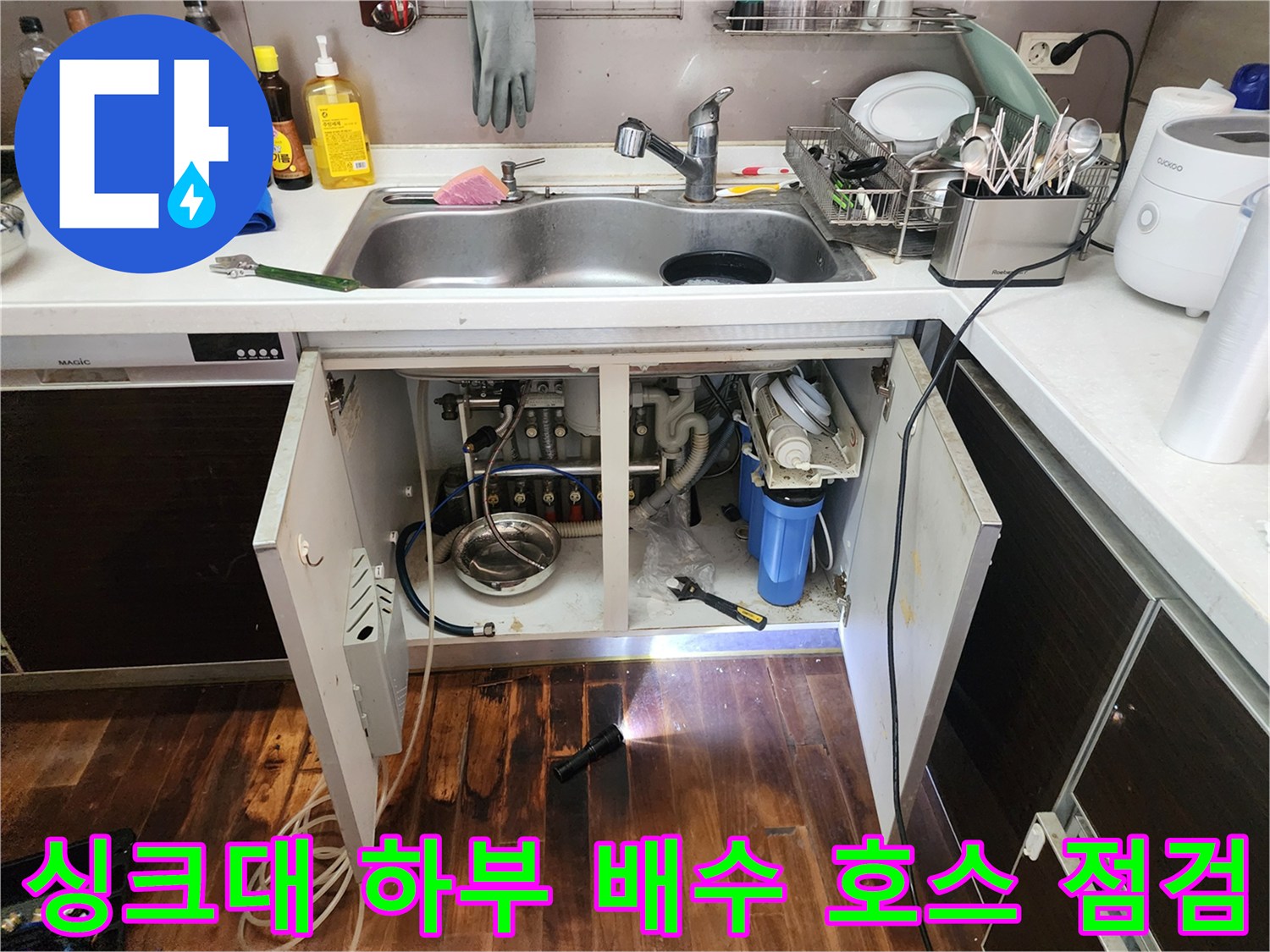 어두운 싱크대 아래쪽 공간을 손전등으로 비추며 배관의 누수 흔적을 찾는 장면