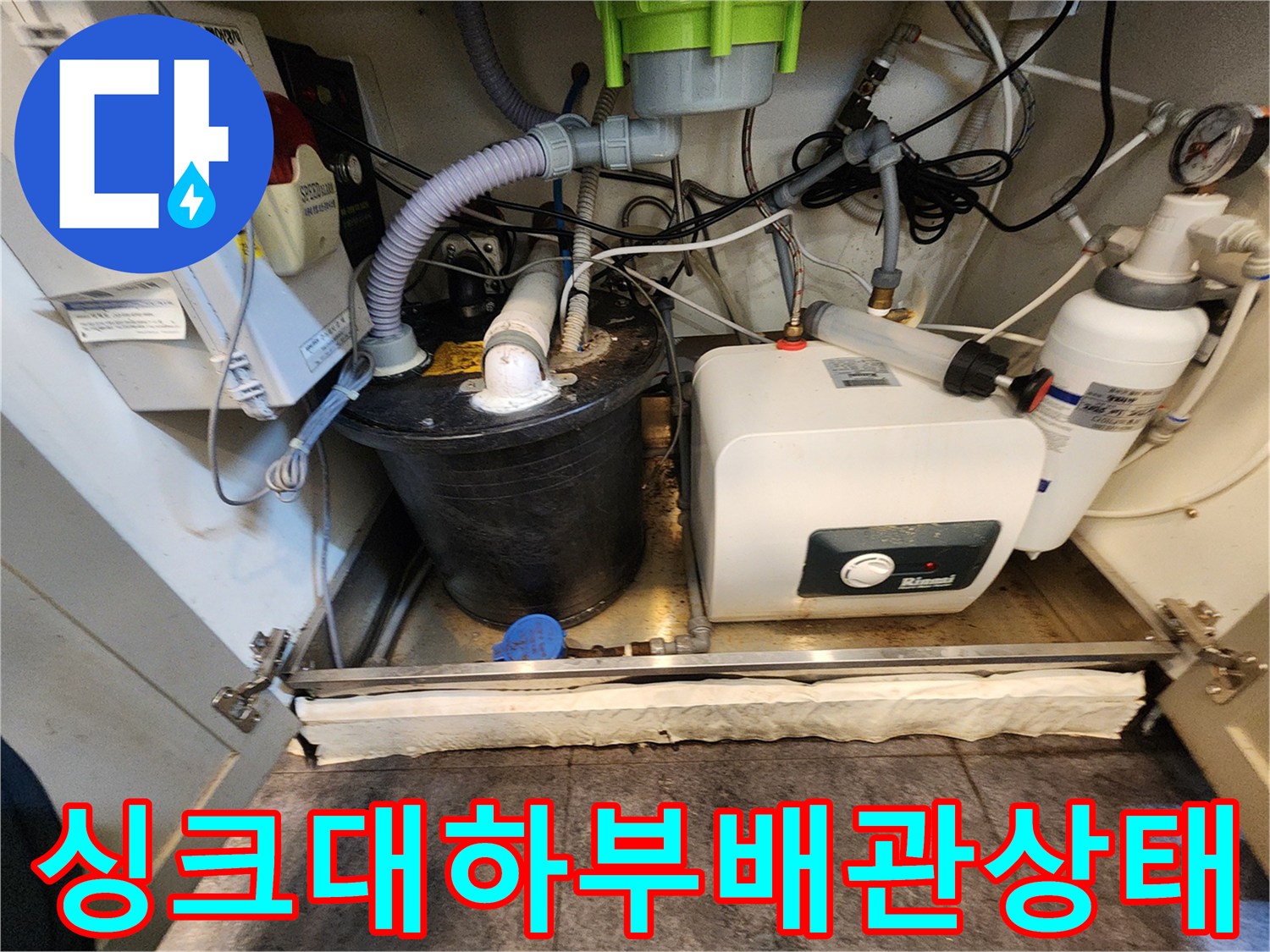 복잡한 배관과 온수기가 설치된 싱크대 하단부 모습입니다.