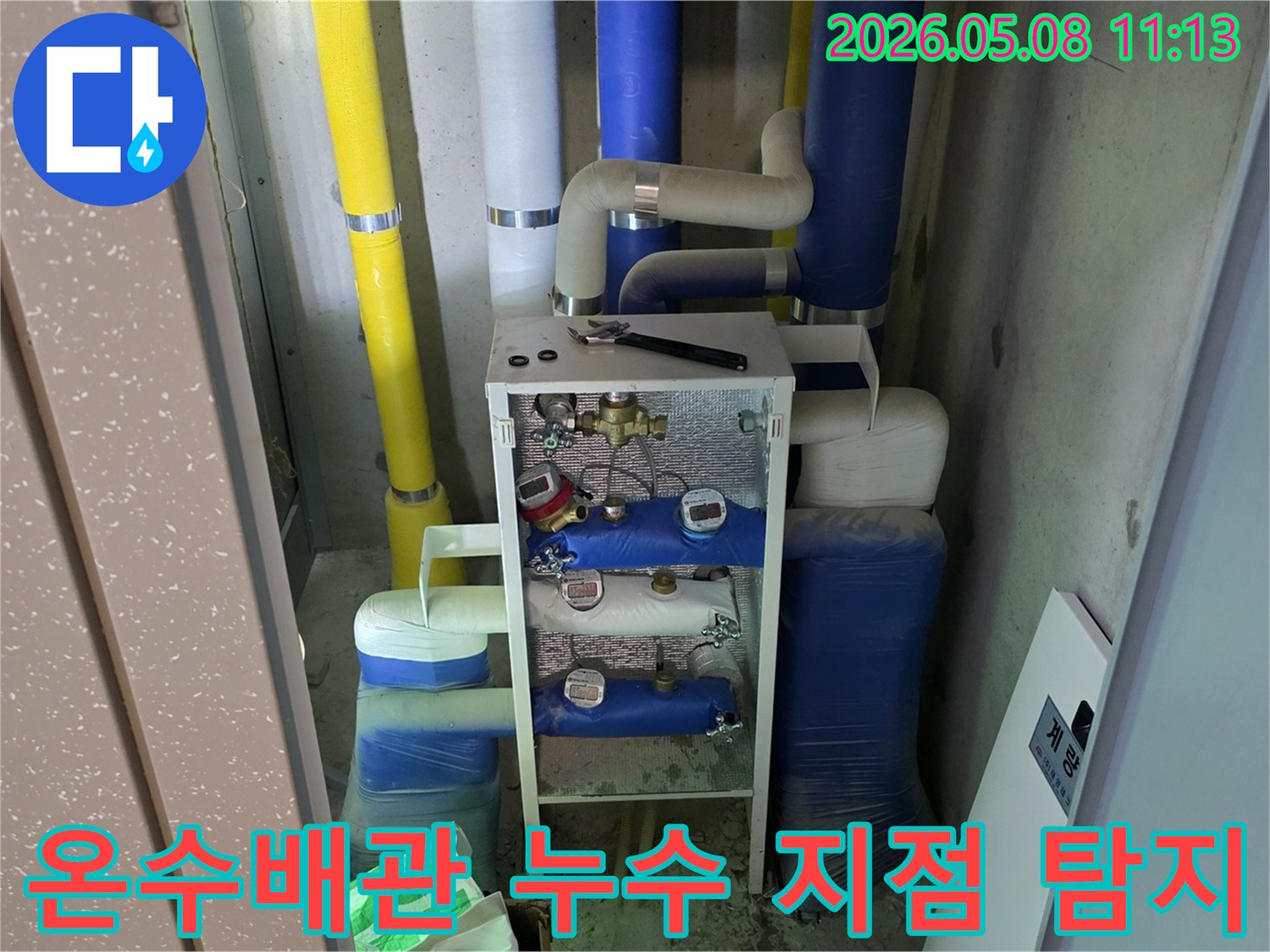 이 사진은 건물의 유틸리티 공간에 설치된-다양한 색상의 단열재로 감싸진 배관들과-수도 계량기-그리고 작업 도구를 보여줍니다-이는 누수 문제 해결을 위한-전문적인 접근 방식을 나타냅니다