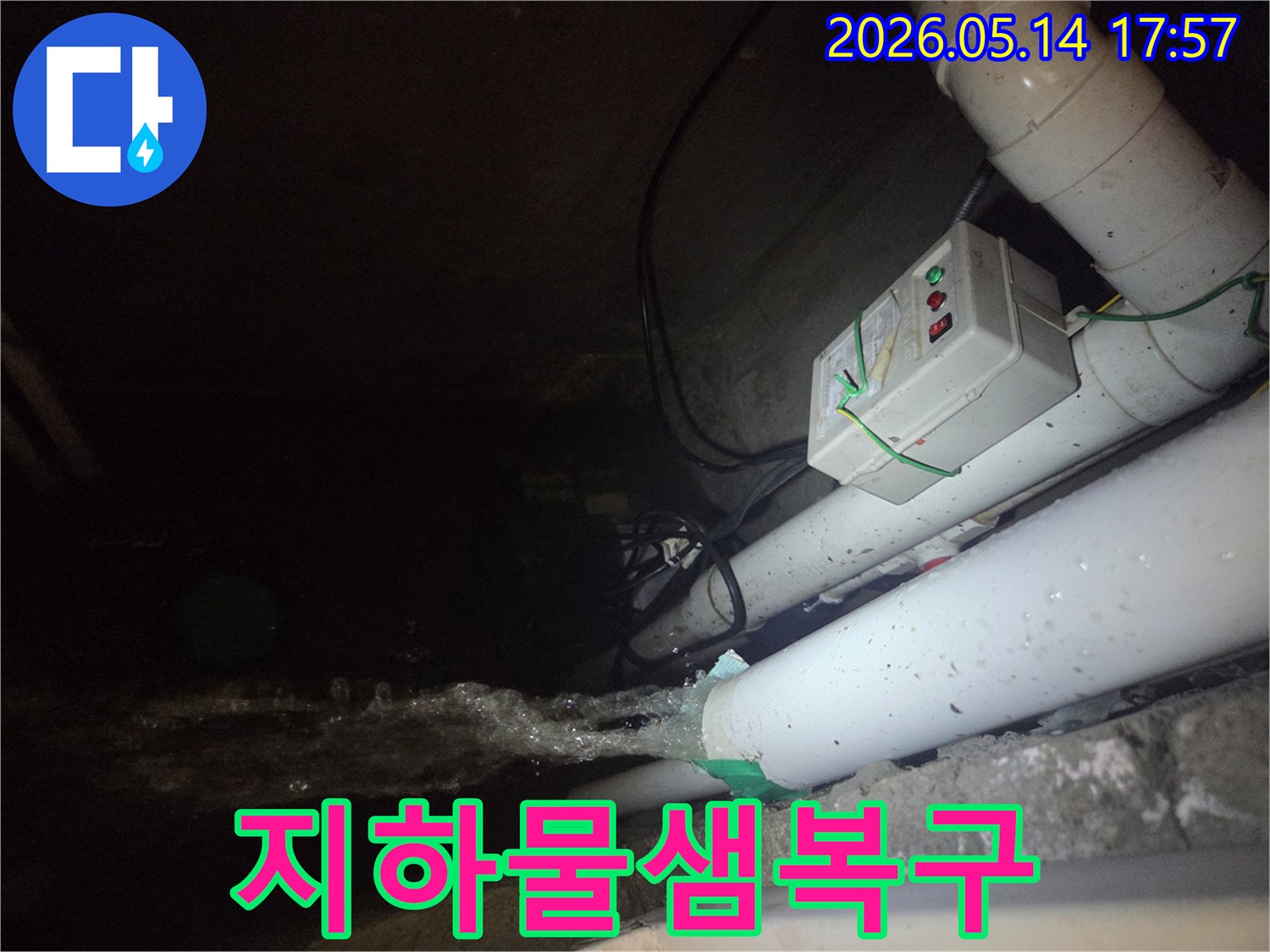 어두운 지하 공간에서-파이프에서 물이 강하게 뿜어져 나오는-주택 지하 물샘 현장을 보여주는 사진입니다-전기 제어 박스와 배관들이 함께 보입니다.