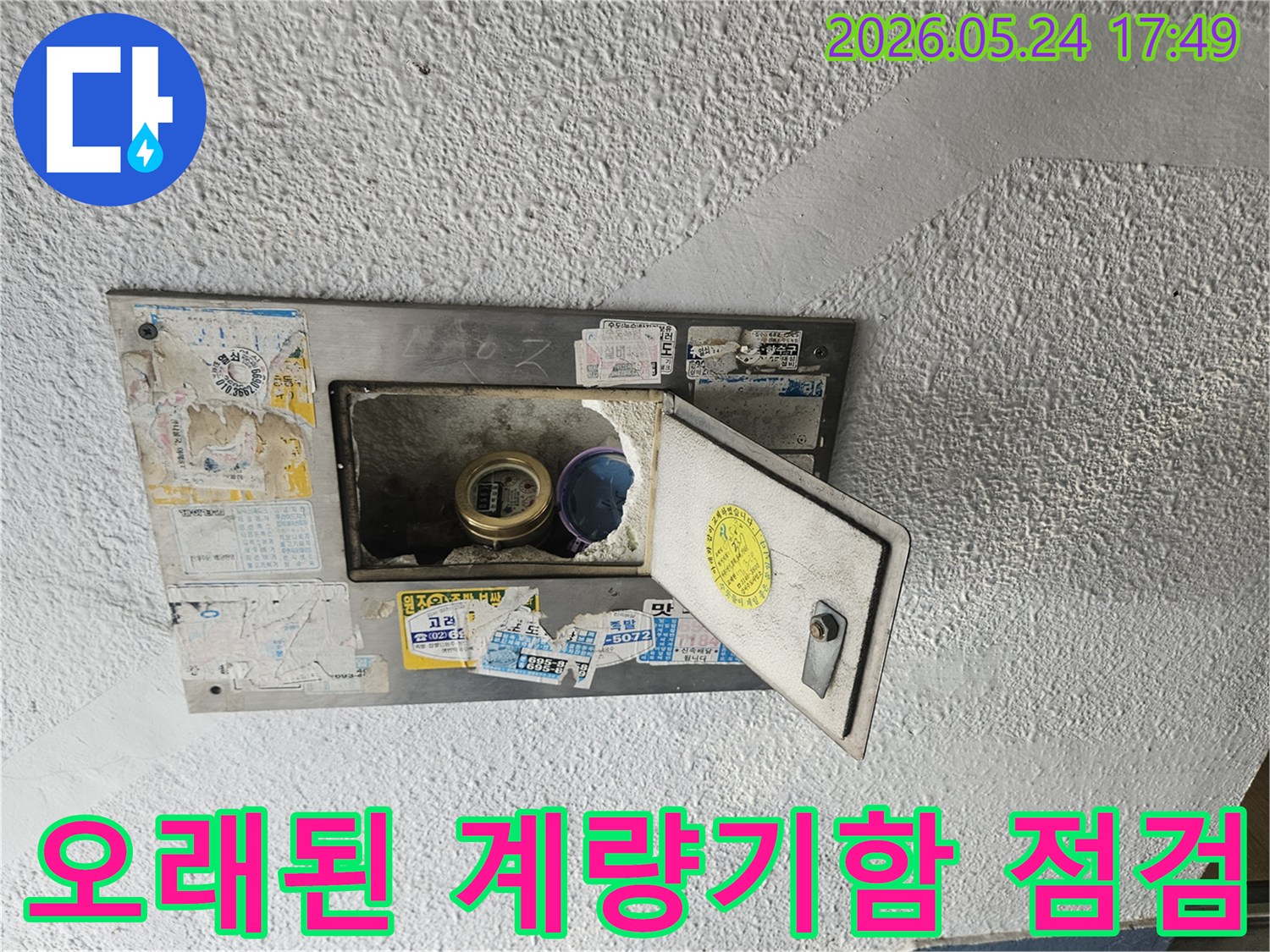 낡고 오래된 수도계량기함이 열려있고-내부의 수도계량기와 주변 상태를-점검하는 모습이 담긴 사진입니다.