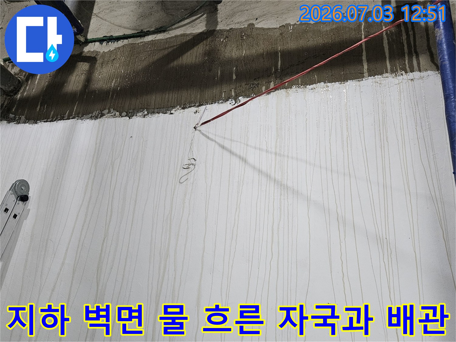 천장의 콘크리트 턱 아래로 하얀 벽면에 수많은 세로형 누수 얼룩이 있고 우측에 파란색 배관들이 배치된 모습
