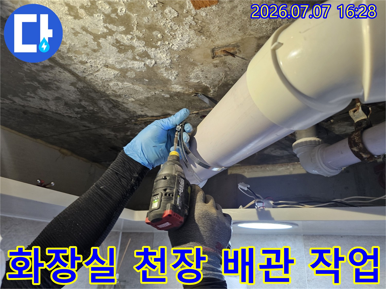 파란 장갑을 낀 엔지니어가 화장실 천장 내부의 노후된 배관 부위를 전동공구로 점검하는 모습