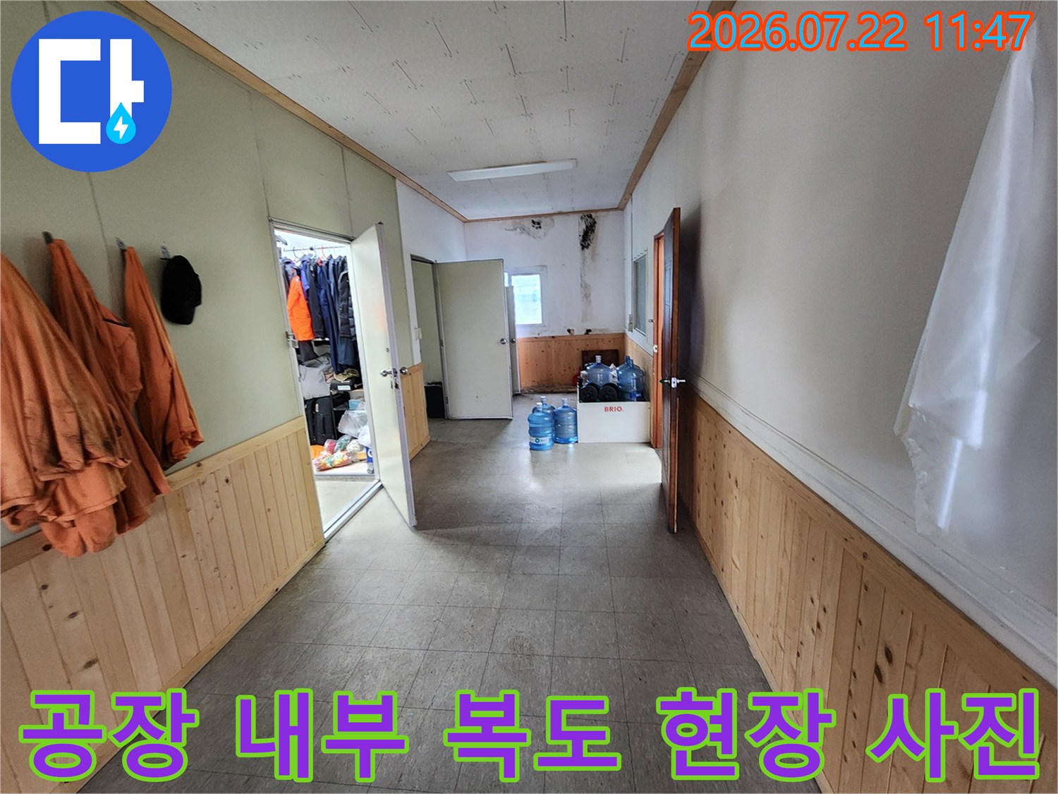 벽면에 주황색 작업복이 걸려 있고 파란색 말통이 놓여 있는 공장 내부 복도 모습