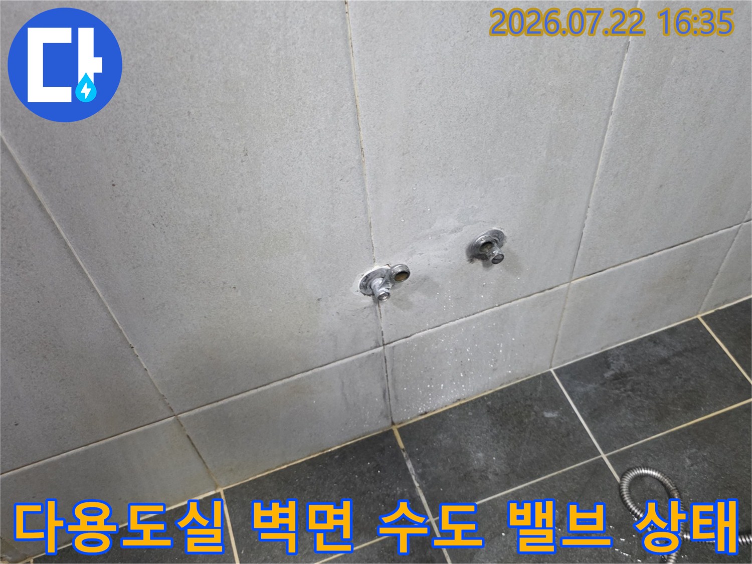 회색 타일 벽면에 설치된 두 개의 수도 밸브와 바닥에 놓인 금속제 호스-누수 점검 현장