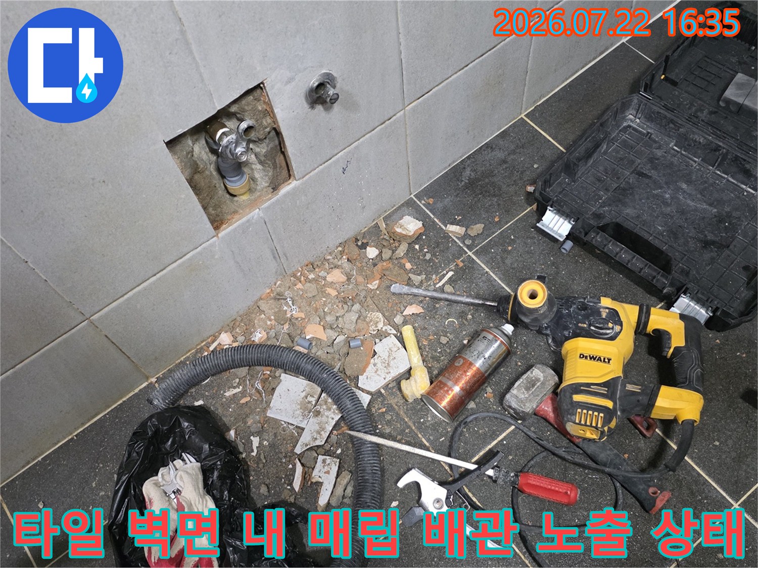 흰색 타일 벽이 사각형으로 뚫려 있고 그 안에 금속 배관이 드러나 있으며 바닥에 공구가 놓인 모습