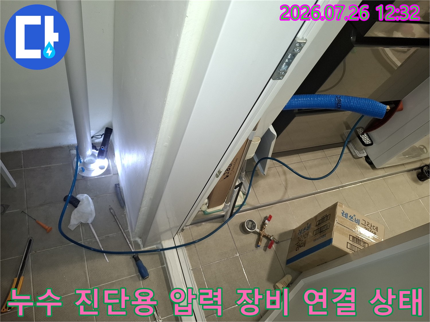 흰색 기둥 옆으로 파란색 배관이 지나가고 바닥에는 누수 테스트 장비와 공구가 놓인 모습