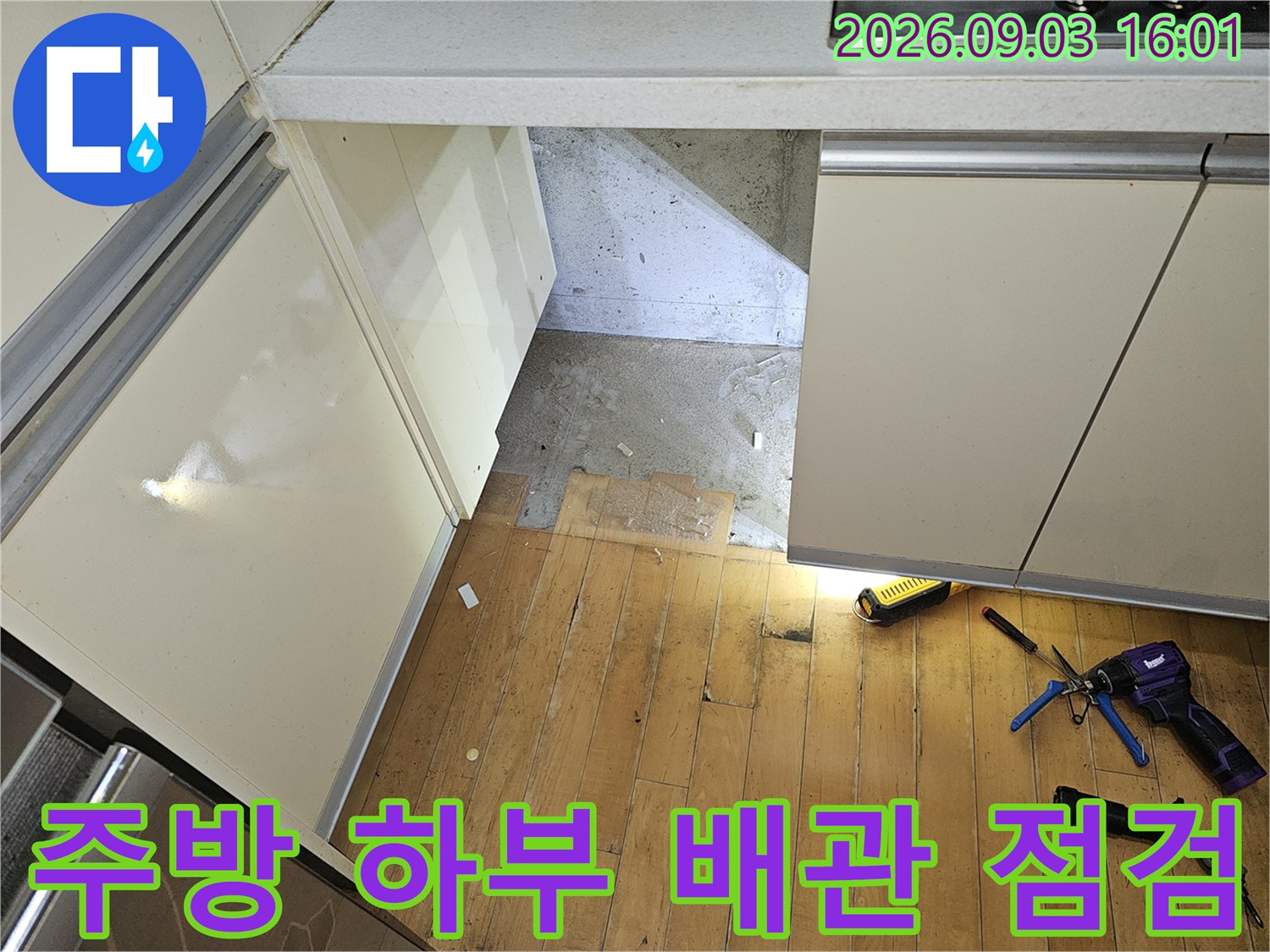 손전등으로 비춘 싱크대 하단 바닥의 콘크리트 노출 부위와 배관 경로