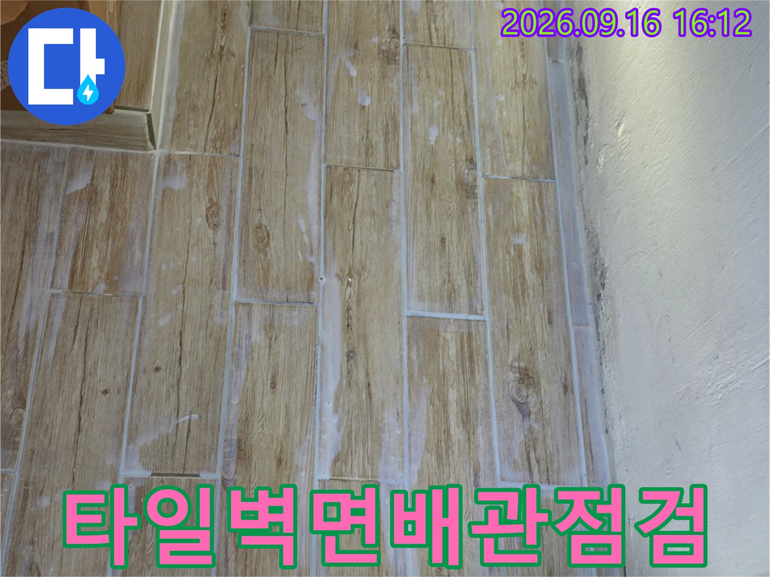 베이지색 타일 벽면에 설치된 흰색 배관과 연결 부위를 점검하는 모습입니다.